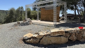 Una estructura metàl·lica amb una plataforma de fusta, envoltada per una paret de pedra i un sòl de grava, amb arbres al fons.