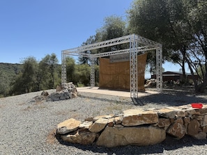 A metal structure with a wooden platform, surrounded by a stone wall and gravel ground, with trees in the background. - Moonshadow Flat Glamping: Quartz Tent (Penn Valley)