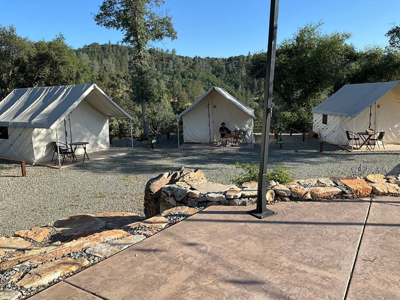 A campsite with three tents, each with a table and chairs, set against a backdrop of trees and hills.