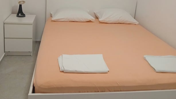 Quarto individual com cama de solteiro, cabeceira branca, mesa de cabeceira com abajur e janela com persianas.
