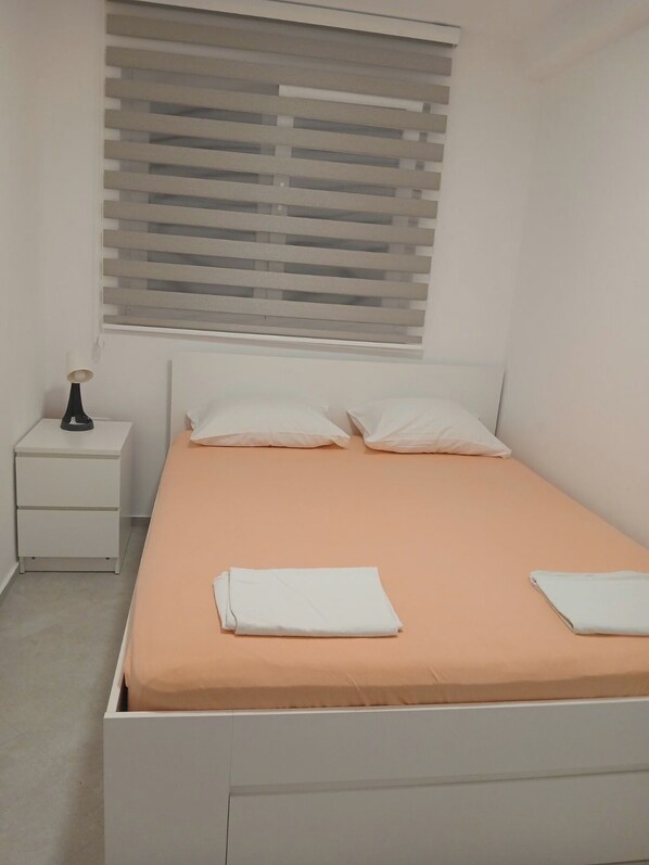 Habitación individual con una cama de soltera, cabecera blanca, mesita de noche con lámpara y ventana con persianas.