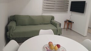 Uma sala de estar com um sofá verde, uma mesa de jantar branca com frutas, uma TV fixada na parede e uma pequena mesa lateral de madeira.