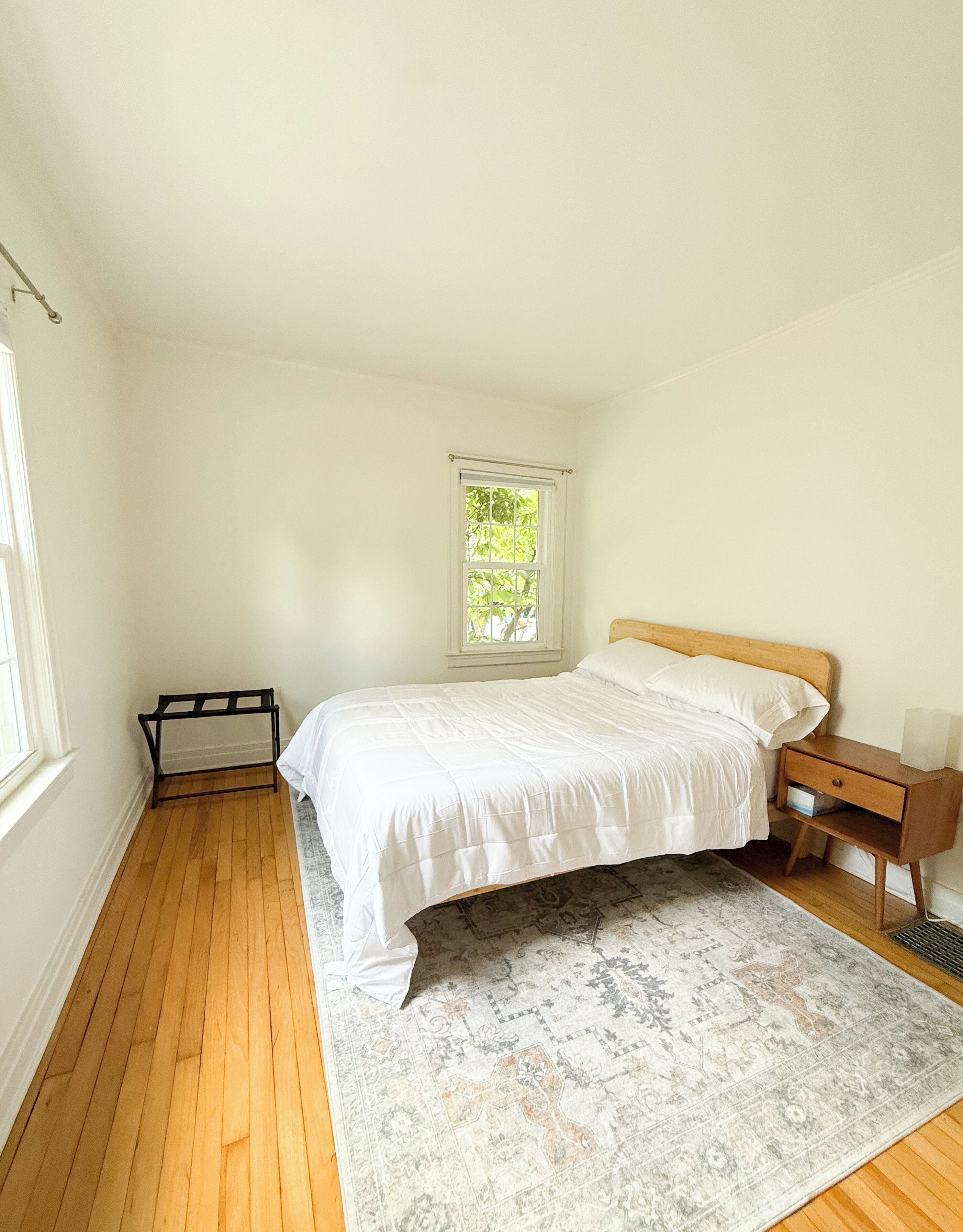 A bedroom with a single bed, a nightstand, a window with a view of greenery, and a patterned rug.