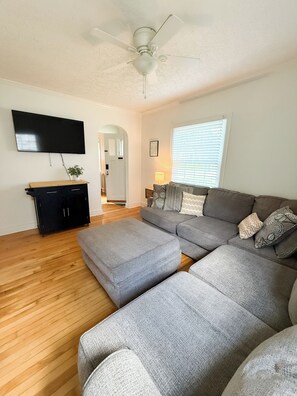 A living room with a grey sectional sofa, a flat-screen TV mounted on the wall, a ceiling fan, and a wooden floor. - Cozy and Convenient - Retreat on Rosemont - Kid Friendly - Covered Back Deck (Lexington)