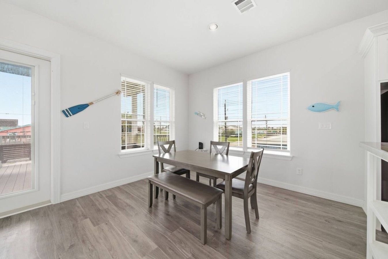 A dining area with a wooden table and chairs, large windows with blinds, and a view of the outside.