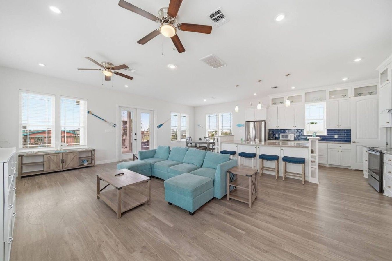 A spacious living area with a sectional sofa, a dining table with bar stools, and a kitchen with white cabinets and blue backsplash.