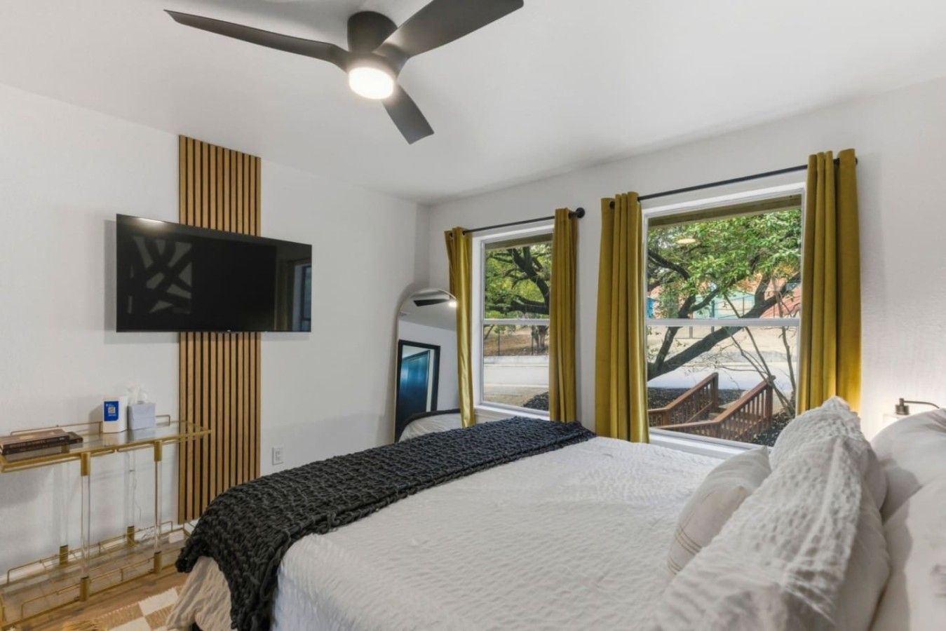 Une chambre à coucher avec un lit, un téléviseur fixé au mur, une fenêtre avec des rideaux et un ventilateur de plafond.