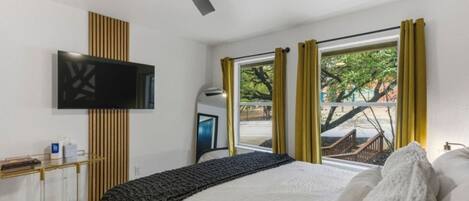 Une chambre à coucher avec un lit, un téléviseur fixé au mur, une fenêtre avec des rideaux et un ventilateur de plafond.
