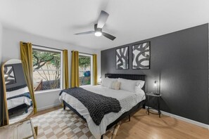 Un dormitorio con una cama, ventilador de techo, ventana con cortinas y una obra de arte en la pared.