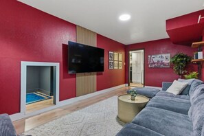 A living room with a red wall, a flat-screen TV, a grey sofa, a small round coffee table, and a door leading to another room.