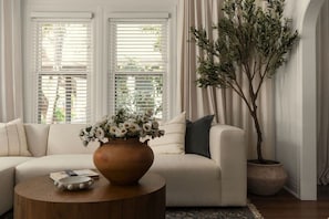 Ein Wohnzimmer mit einem beigen Sofa, einem runden Holztisch, einer Vase mit weißen Blumen, einer Topfpflanze und einem Fenster mit Jalousien.