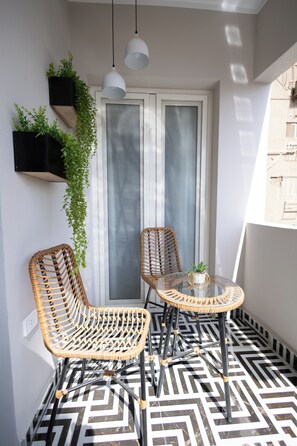 Ein Balkon mit zwei Korbstühlen, einem Glastisch, hängenden Pflanzen und Blick auf ein Gebäude durch eine Milchglasschiebetür.