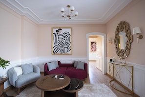 Quarto com sofá vermelho, poltrona cinza, mesa de centro redonda de madeira e uma grande pintura abstrata na parede.