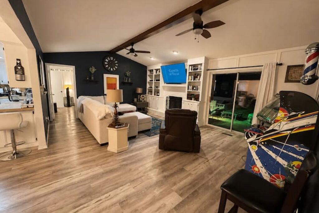 A living room with a sofa, armchair, and a television. There is a kitchen area with a bar stool and a dining table. The room has wooden flooring and a ceiling fan.