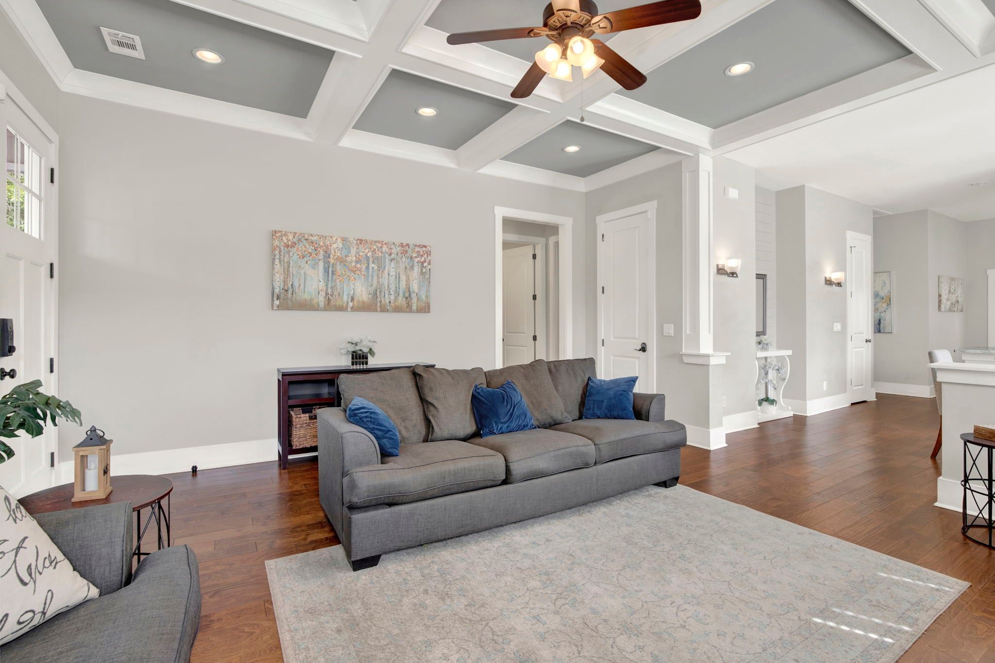 A spacious living room with a grey sofa, a wooden ceiling fan, and a large painting on the wall.