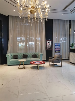 A hotel lobby with a tufted light blue sofa, a round coffee table with a Captain America shield, a grey armchair, a chandelier, and a large window with sheer curtains. - Xana Hotelle (Guangzhou)