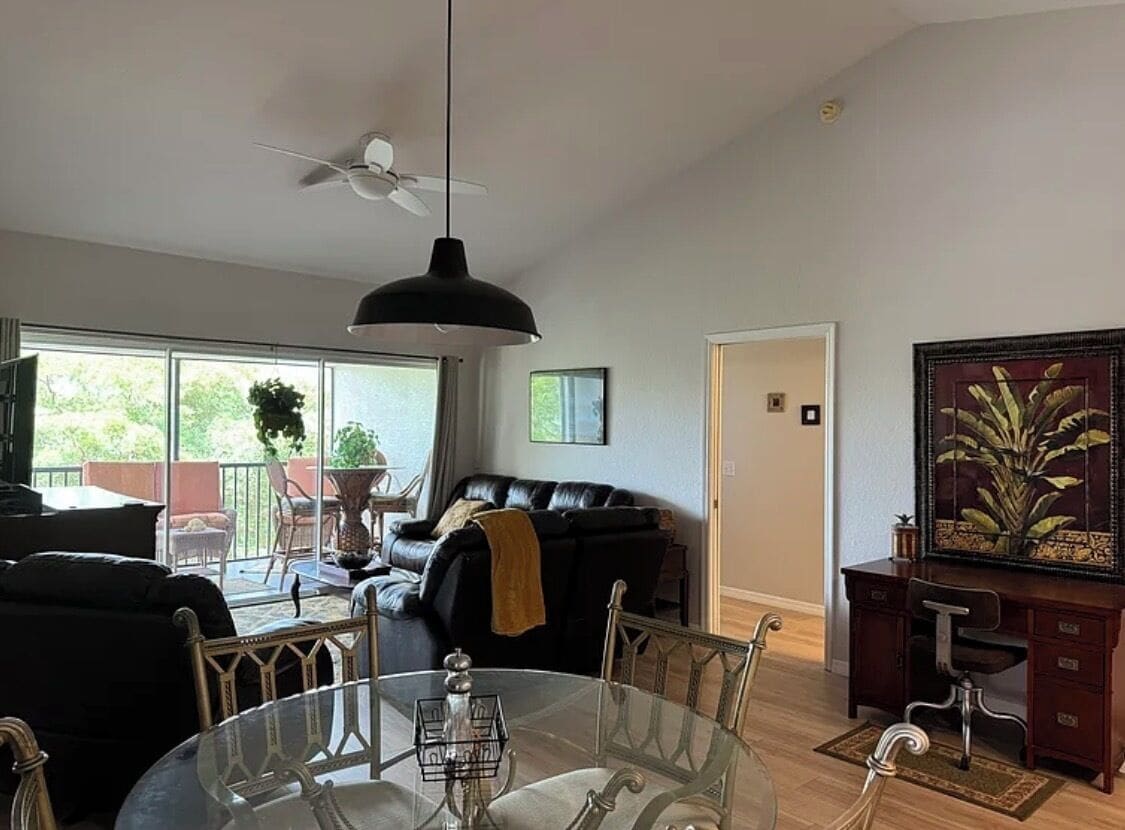 A living room with a glass dining table, a black sofa, a ceiling fan, and a painting on the wall.