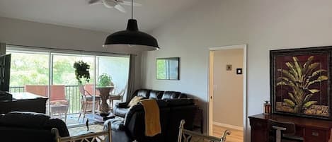 A living room with a glass dining table, a black sofa, a ceiling fan, and a painting on the wall.