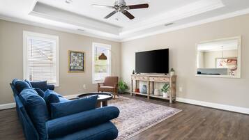 Un salon avec un canapé bleu, un téléviseur, un ventilateur de plafond et une grande fenêtre équipée de stores.