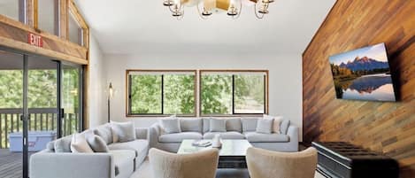A modern living room with a sectional sofa, two armchairs, a coffee table, and a fireplace. There are large windows providing natural light and a view of the outdoors.