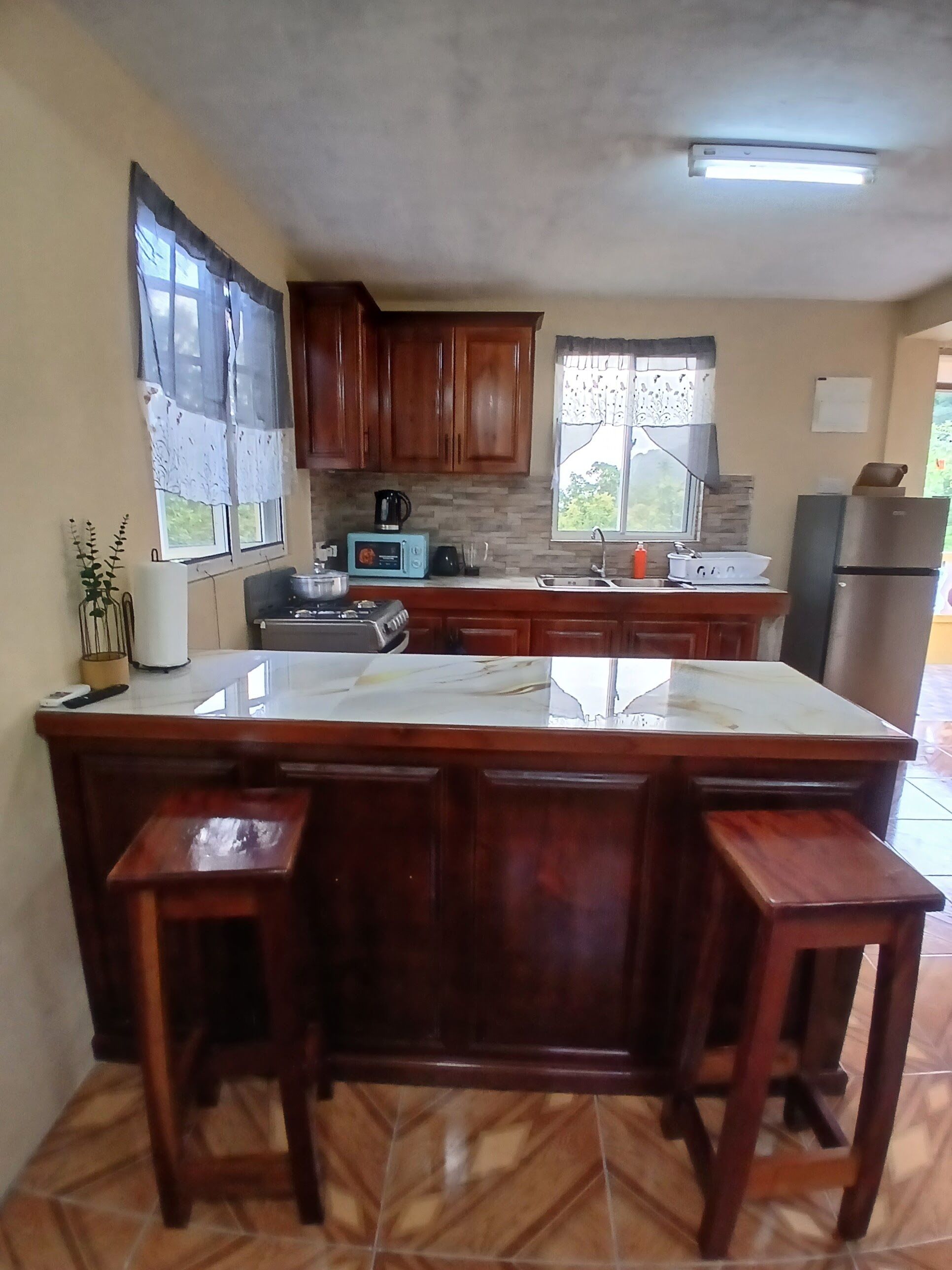 A kitchen with wooden cabinets, a large island with stools, a microwave, and a refrigerator.