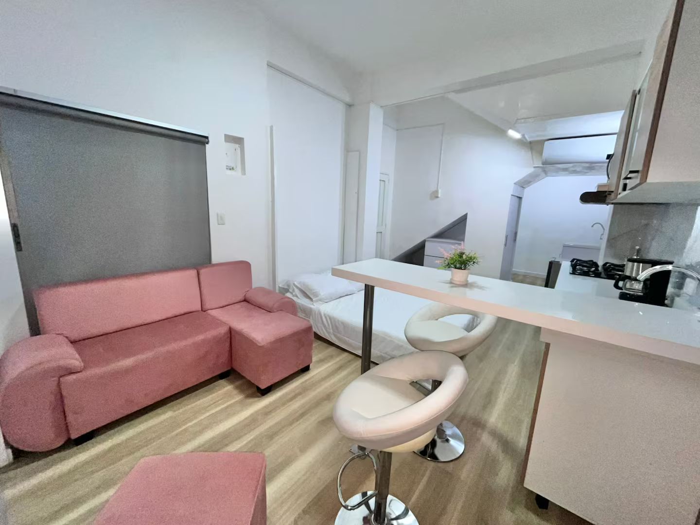 A compact living space with a sofa, a small dining area with stools, a kitchenette, and a bed.