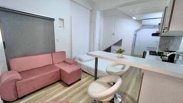 A compact living space with a sofa, a small dining area with stools, a kitchenette, and a bed.