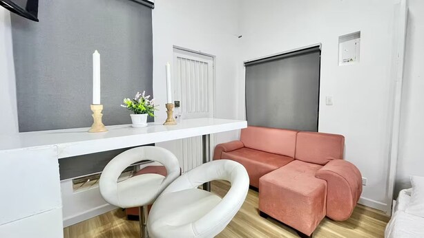 Una habitación con un sofá blanco, dos sillas blancas, una mesa blanca con velas y una planta, y una puerta cerrada.