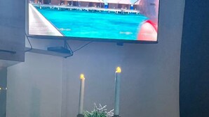 Una habitación con una televisión de pantalla plana montada en la pared, dos velas encendidas sobre una mesa y un par de sillas modernas.