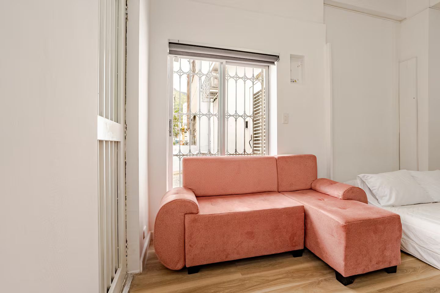 A room with a pink sectional sofa, a bed, and a window with metal bars.