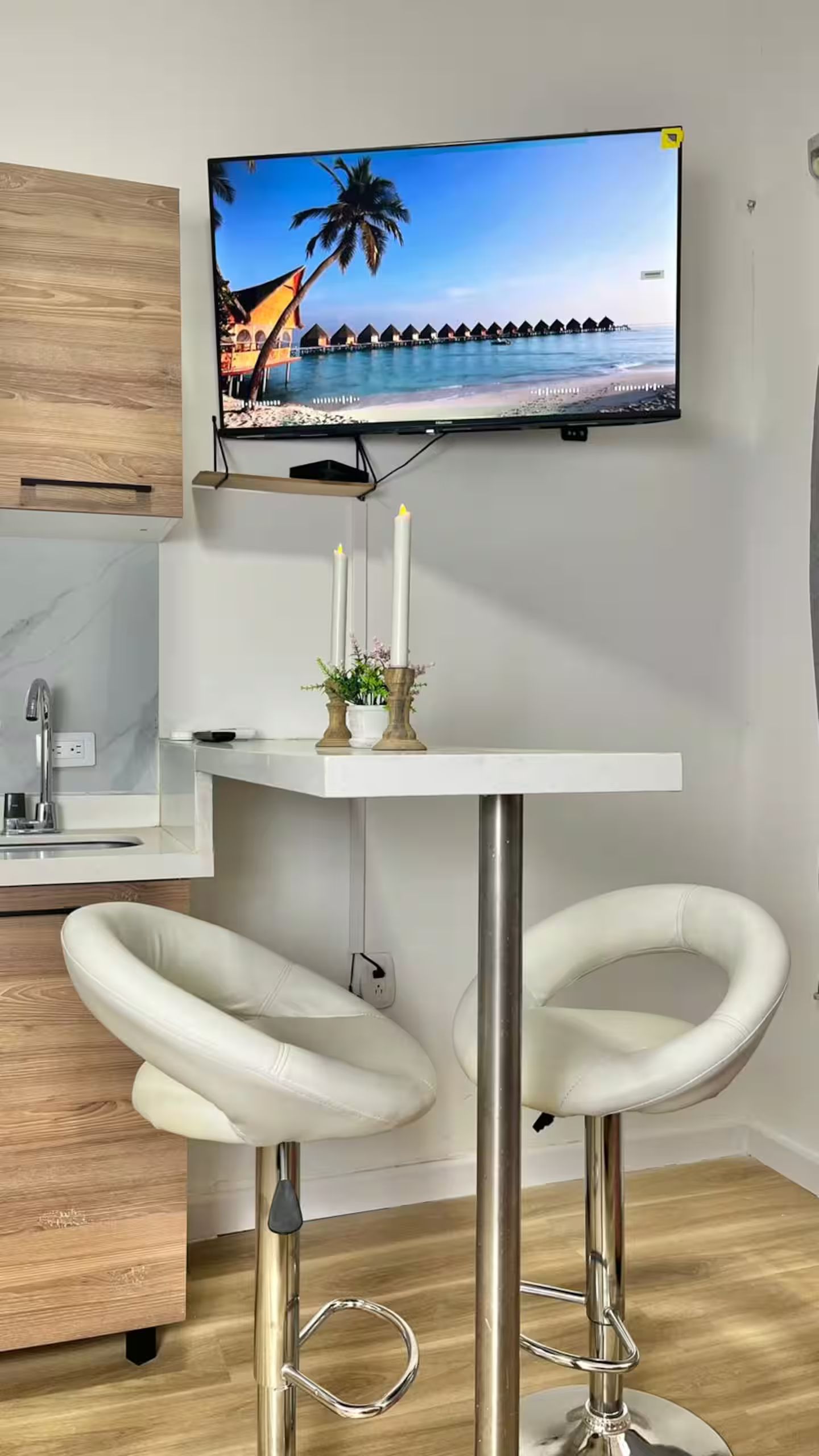 A modern kitchen with a flat-screen TV mounted above a white bar stool area, a marble countertop, and a stainless steel sink.