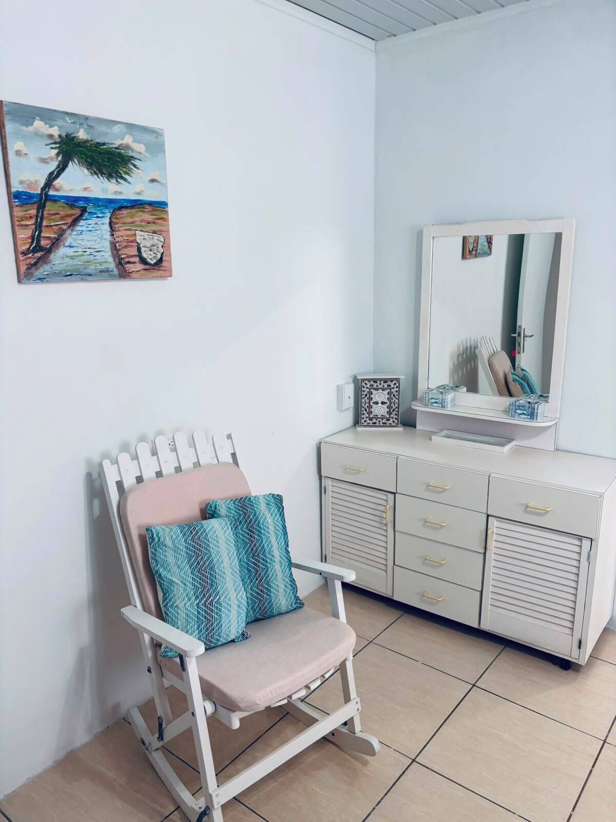 Une pièce avec une coiffeuse blanche, une chaise avec des coussins, un tableau représentant un palmier et un sol carrelé.