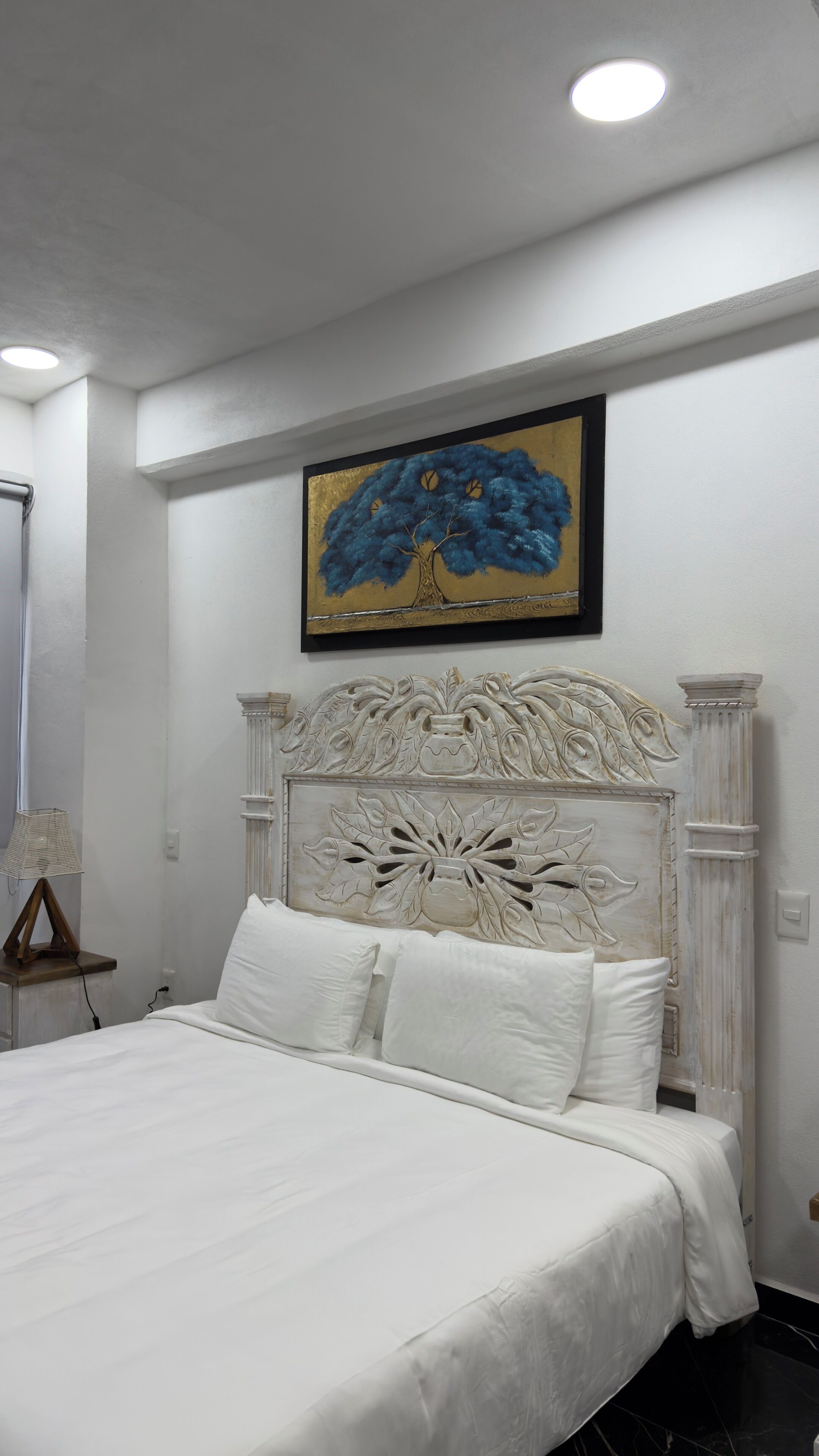 A neatly made bed with white linens, a decorative headboard with carved details, and a painting of a tree on the wall above.