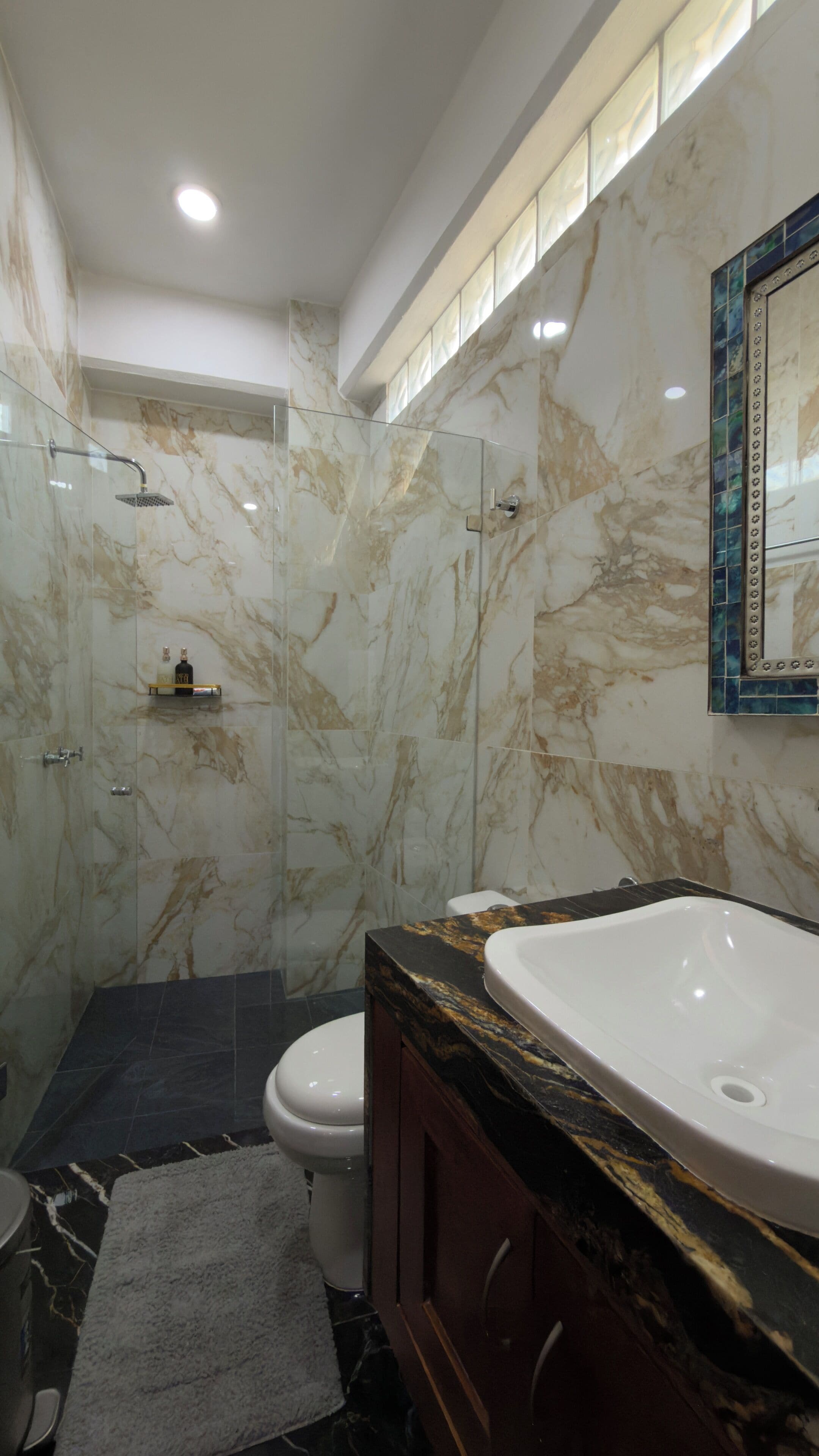 Un baño moderno con ducha envuelta en vidrio, un lavamanos blanco y un revestimiento de mármol en la pared.