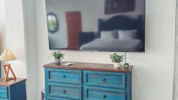 Een kamer met een grote flatscreen televisie aan de muur, een blauwe houten dressoir met meerdere laden, een kleine plant in een pot op het dressoir, en een betegelde vloer.