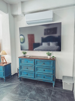 Habitación con una amplia televisión de pantalla plana montada en la pared, un aparador de madera azul con varios cajones, una pequeña planta en maceta sobre el aparador y suelo de baldosas.