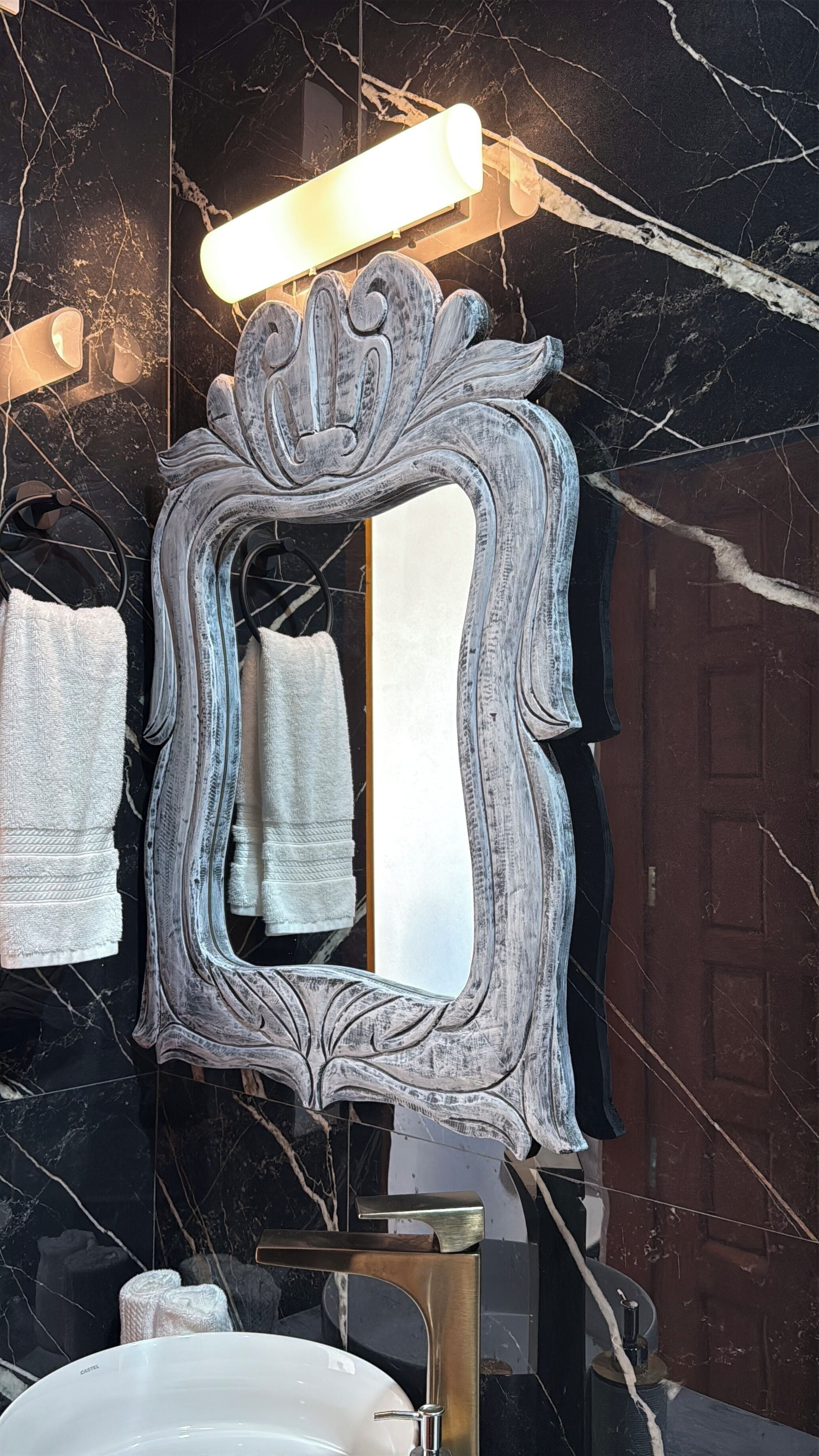 A bathroom with a marble wall, a white sink, a mirror with a decorative frame, and a towel rack with towels.
