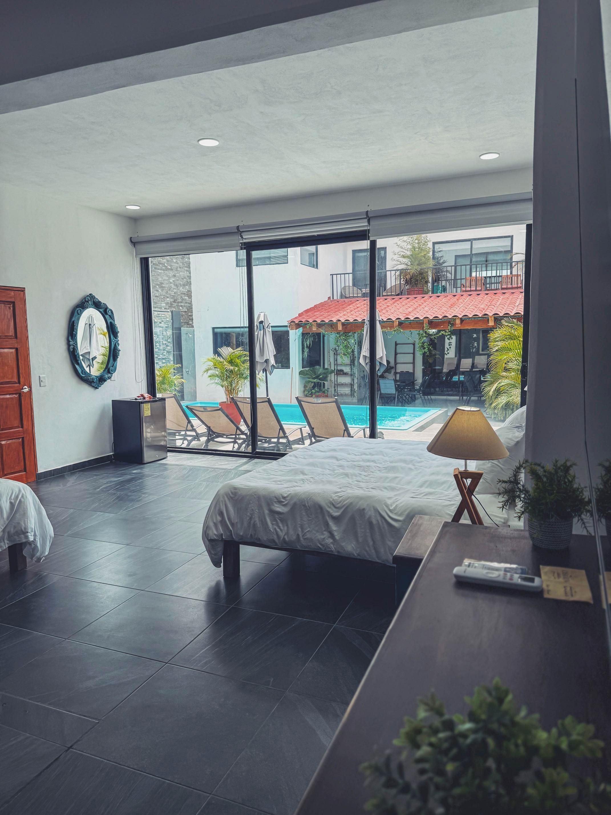 A modern hotel room with a large bed, a desk, a chair, and a view of a swimming pool and outdoor seating area.