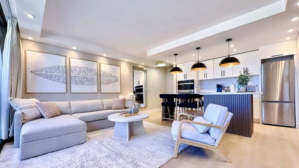Ein modernes Wohnzimmer mit einer Couch, Sesseln und einem Couchtisch. Es gibt eine Küchenzeile mit einem Kühlschrank und Schränken.