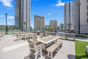 An outdoor patio area with tables and chairs, a barbecue grill, and a tennis court in the background. - 29F-Upscale-Fully Renovated-Waikiki Banyan 1 Bedroom-Ocean View w/ Free Parking (Honolulu)