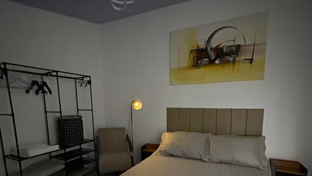 A bedroom with a bed, a chair, a shelf, and a painting on the wall. - Studio 5 | Vila Granados (Embu das Artes)