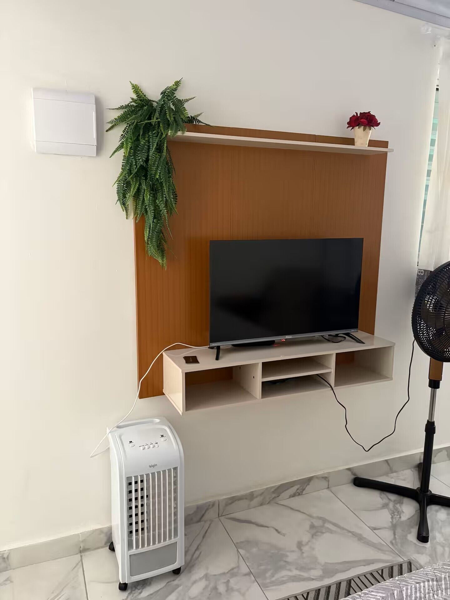 A living room with a flat-screen TV mounted on a wooden wall, a white radiator, a standing fan, and a potted plant.