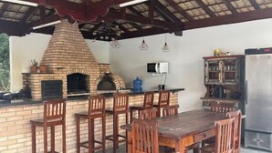Ein Außenbereich mit Küche, der ein Backrohr aus Ziegeln, Holzsessel, einen Esstisch und einen Kühlschrank umfasst.