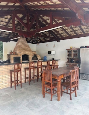 An outdoor kitchen area with a brick oven, wooden chairs, a dining table, and a refrigerator. - Vrbo Property (Nazaré Paulista)