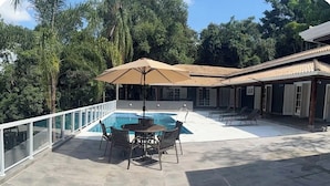 Ein Poolbereich mit Tisch und Stühlen, umgeben von einer Glasbrüstung, und ein überdachter Sitzbereich mit Sonnenschirmen.