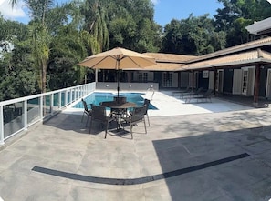 A pool area with a table and chairs, surrounded by a glass railing, and a covered seating area with umbrellas. - Vrbo Property (Nazaré Paulista)