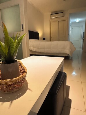 A hotel room with a bed, a bedside table with a potted plant, an air conditioning unit, and a door leading to another room.