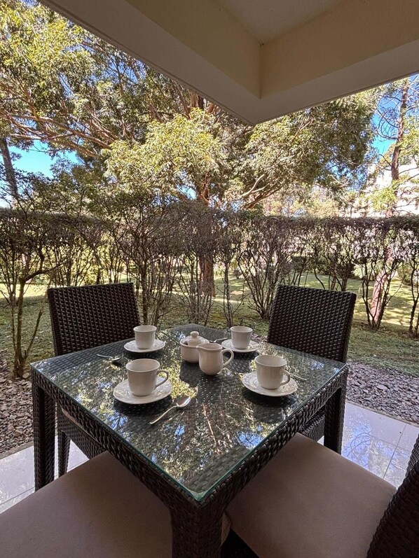 An outdoor dining area with a glass-top table, wicker chairs, and a view of trees and grass.