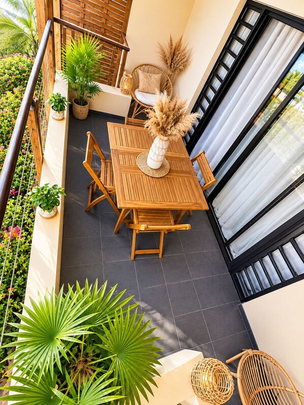 A balcony with a wooden table and chairs, surrounded by potted plants and a wicker chair.