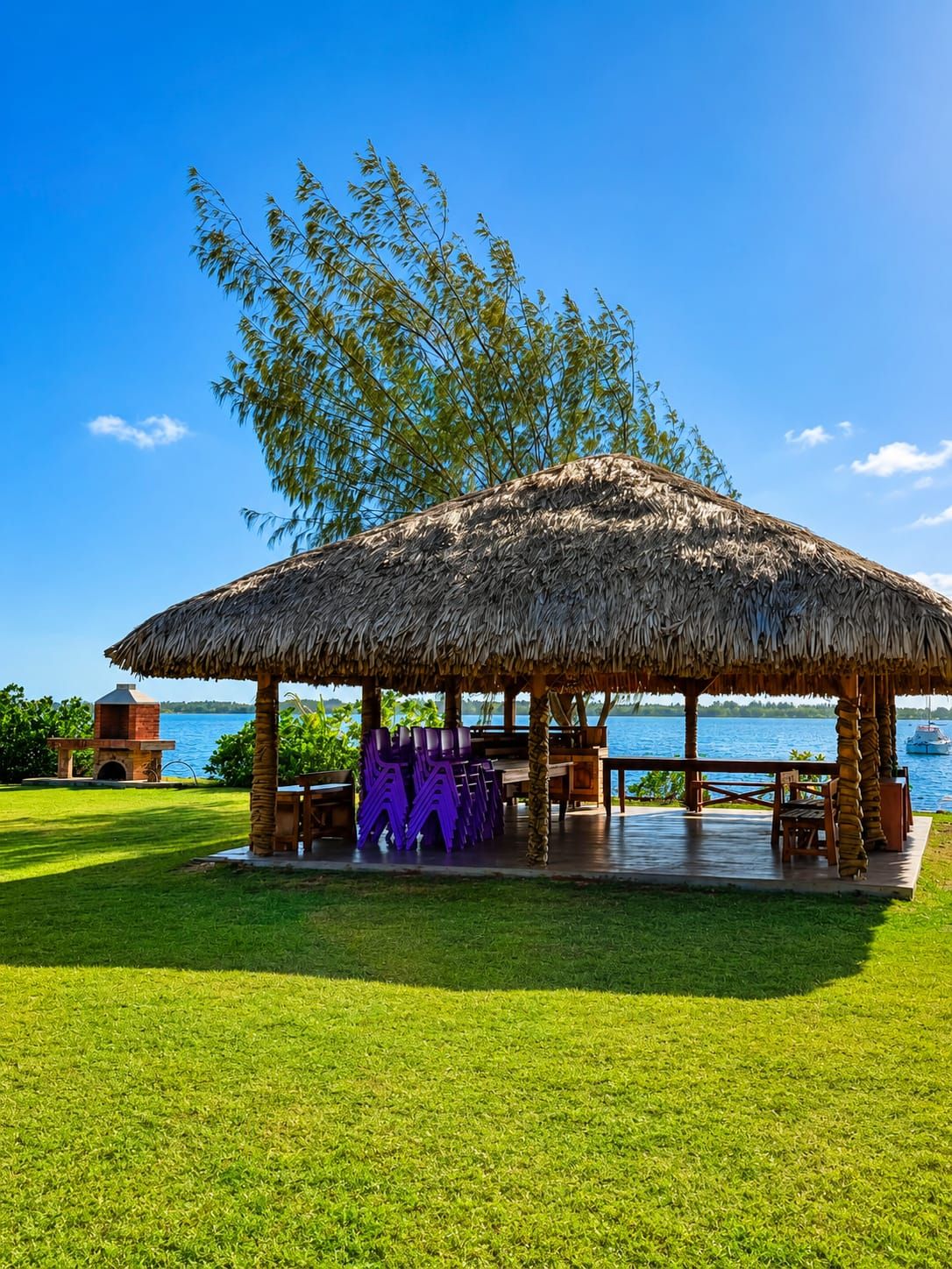 A thatched-roof pavilion with outdoor seating, a grill, and a view of a body of water.